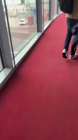 From the perspective of an observer walking alongside a window at an airport, the video captures the external airport environment. The window frames, colored in a light grey or white, create sections displaying the outdoors. The airport tarmac is visible through the windows, displaying vehicles such as buses and service trucks moving about. A service truck with red accents moves on the tarmac. The ground is a dark grey color, possibly indicating dampness, and is demarcated with white lines and geometric shapes. In the background, airport buildings can be seen, contributing to the overall atmosphere. Inside, the flooring is covered with a vivid red carpet. The video's focus remains on the external activity and the airport environment, with the red carpet providing a stark contrast to the industrial scenery outside.