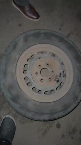 A tire on a steel rim is on the pavement. The camera moves to reveal the legs and feet of several people and the undercarriage of a vehicle. One person is working on the vehicle.