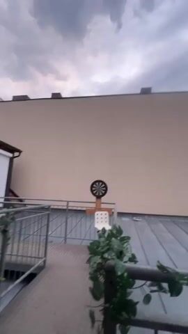 The video shows a dartboard mounted on a wooden stand outdoors. It is positioned on a metal balcony railing with artificial green plants draped over it. In the background, there is a light-colored building with a gray metal roof. The sky is overcast with dark clouds. The camera pans to the right, revealing more of the building's roof and a small satellite dish. There are no people or animals visible in the video.