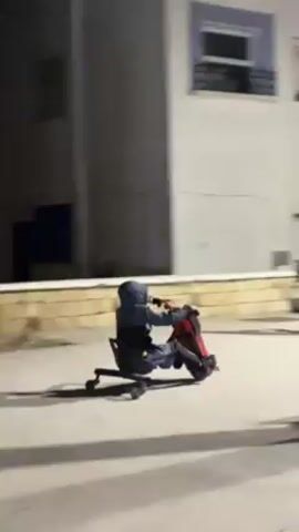 The video shows a person riding a modified office chair down a paved area at night. The person is wearing a hooded sweatshirt and appears to be propelling the chair with their hands or feet. In the background, there is a building with lit windows, a street light, palm trees, and a vehicle covered in brightly colored lights. The person is moving away from the camera. The video appears to be filmed from a handheld device.