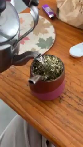 The video shows a silver teapot pouring hot water into a brown and pink mate cup filled with yerba mate. The cup has a silver straw (bombilla) sticking out of it. The cup is sitting on a wooden table, along with a brown coaster, a lighter, a bag, and a white remote control. A person's legs are visible in the bottom left corner of the frame.