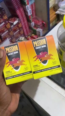 The media file shows a person's hand holding two boxes of "PASSION ENERGY DRINK". The boxes are bright yellow with orange and red accents. The design features silhouettes of horses with riders in a race. Text on the boxes indicates "NO PRESERVATIVES" and "SUGAR FREE". Below this, it lists "NAFDAC Reg. No. 01-9933" and "FDB/DR/03-126". It also states "Content: 6 sachets @ 4g". The background appears to be a retail setting, possibly a store or market stall, with shelves of products visible. Some of these products are stacked and appear to be of various sizes and colors, including what look like more drink boxes or packets. A clear plastic jar with a yellow lid is also visible on the right side. The lighting suggests it could be daytime, but no specific time is discernible. The scene is likely indoors. The person holding the boxes is not fully visible, but their hand indicates they are likely an adult. There are no discernible emotions conveyed. The location context is Jere South, Nigeria.