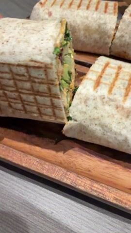 The video shows a wooden serving platter with four cut wraps on it.  One wrap is being lifted up to show the filling.  The wraps are made with a light brown tortilla and have grill marks.  The filling includes what appears to be shredded lettuce, spinach, quinoa and a sauce. A person's hands and lower legs are seen in the background, suggesting the platter is sitting on a table in front of them.