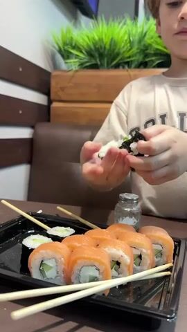 A young child with curly blonde hair, wearing a light-colored t-shirt with the word "COLLEGE" printed on it, is sitting at a table and preparing sushi rolls. The child's hands are visible as they carefully assemble a sushi roll. In front of the child is a black plastic container filled with prepared sushi rolls. Some of the rolls are topped with salmon and cream cheese, while others appear to be cucumber rolls. Several pairs of wooden chopsticks are placed on top of the sushi. The background of the scene shows a wooden wall and some greenery, possibly a potted plant. The lighting suggests an indoor setting.