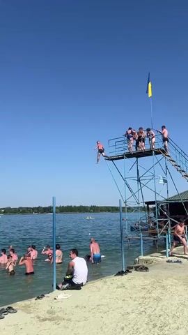 A person jumps from a high dive into a lake. The diver is mid-air and appears to be performing a trick. Several other people are in the water below, watching the jump. A group of people are standing on the diving platform, waiting their turn to jump. The sky is clear and blue, and the sun is shining. The water is a deep blue color, and there are trees in the background. The overall atmosphere is one of fun and recreation on a summer day.