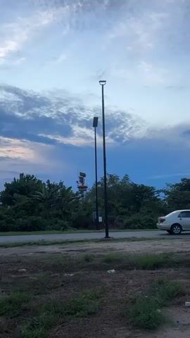 The video shows a landscape with two lampposts in the foreground and lush green vegetation in the background. A road runs horizontally through the scene. In the far distance, a red and white tower-like structure rises above the trees. The sky is a mix of blue and gray with scattered clouds. The ground near the camera is a combination of dirt and grass.