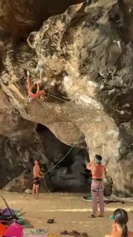 A person is rock climbing on a light-colored rock formation that overhangs a sandy beach. The climber is suspended by ropes and using handholds. Below, on the beach, another person is holding the rope, providing support. Nearby, two other people are watching the climber; one is holding up a device to seemingly capture photos or video. The environment is a beach setting with a rocky overhang, suggesting a coastal or tropical location. Scattered on the sand are various personal items, including a colorful bag and a pair of shoes.