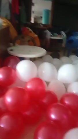 The video begins with a view of red and white balloons scattered across the floor of a room. A woman is sitting on a couch in the background, looking at her phone. The camera shifts to a view of the ceiling and then to a group of people, many wearing red shirts, gathered in a room with a large window. Some of them are holding white balloons in front of their faces. A woman in the center of the group is laughing and obscuring her face with her hands.