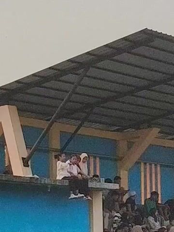 A low-angle, medium shot captures a group of people sitting on a concrete ledge under the roof of a stadium. Three young people, two females and one male, are in the foreground, sitting closely together on the ledge. The two females are wearing hijabs, one light brown and one white, and a white shirt. The male is wearing a white shirt and dark pants. They appear to be watching an event happening below. Below them, a crowd of spectators is visible, seated in tiered seating. The roof above is made of corrugated metal, supported by metal beams. The overall lighting suggests it is daytime, likely late afternoon or early evening, with a slightly overcast sky. The environment appears to be a sports venue.