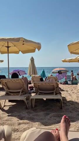 The video shows a beach scene from the perspective of someone lying on a lounge chair. In the foreground, the legs and feet of the person are visible, with painted toenails. Beyond the person, several empty lounge chairs are arranged on the sand, some covered by large, pale yellow umbrellas. In the mid-ground, other beachgoers are seen relaxing on lounge chairs or standing near the water. Various colorful umbrellas, including a striped one and a teal one, are visible. The sea stretches out to the horizon under a clear blue sky. The sand appears to be a mix of small pebbles and fine grains. People are dressed in beachwear, and the overall atmosphere suggests a sunny day at the beach.