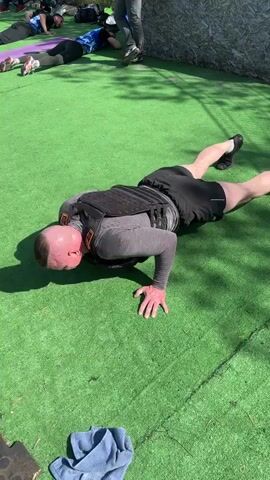 A group of people are doing push ups on a green surface that resembles artificial grass. The focus shifts between two men wearing weighted vests who are in a push-up position. They both have fair skin and are wearing athletic clothes. The first man is wearing a grey long sleeve shirt and black shorts. The second man is wearing a yellow tank top and yellow shorts. He is asked in Russian if he has a plan he is sticking to. A grey towel lies on the ground near the first man. Other people can be seen lying on the artificial grass in the background. A section of a wooden fence can be seen along the edge of the artificial grass.