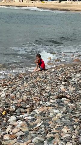A young child wearing a red and blue long-sleeved shirt and blue shorts is seated on a rocky beach, facing the ocean. Small waves gently lap at the child's feet as they play with the water. The beach in the foreground is covered in various sizes and colors of pebbles and stones. In the background, across a body of water, a sandy beach with several buildings, some trees, and a few distant people are visible under a clear sky.