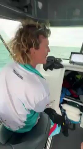 The video starts with a person standing at the helm of a boat. They are wearing a light blue shirt and patterned shorts. The cabin is visible, with various items stowed inside. The camera then moves to the stern of the boat, showing the wake as it speeds across the water. The sky is overcast and a distant shoreline is visible.