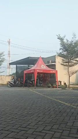 The video pans across an outdoor scene. In the foreground, a paved area with a brick pattern is visible. To the left, a red tent is set up with motorcycles parked nearby. Several people are gathered under or around the tent. Further back, a building structure is present, with a tall communication tower in the distance. To the right, a tree with sparse leaves stands near the building. The sky is overcast and hazy. As the video pans right, more motorcycles are seen, followed by cars parked in a row. One car has a decorative pattern on its side. Electrical poles and wires are also visible in the background. The overall impression is that of a parking lot or an outdoor event area.