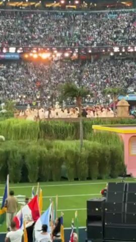 The video shows a performance at a stadium filled with people. In the foreground, there are several people holding flags, and large speakers are visible. In the midground, there is a stage decorated with artificial grass, palm trees, and pink structures. On the stage, a group of performers is dressed in what appears to be white robes. The stadium is packed with a large crowd, and bright stage lights illuminate the scene. The video seems to capture a live musical or cultural performance during a large event.