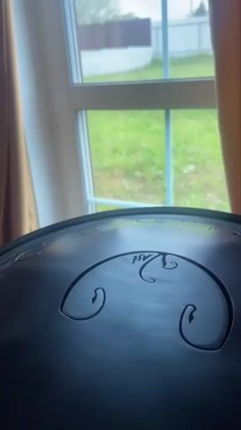 A close-up shot shows a person playing a handpan instrument indoors. The instrument is dark gray and has cut-out shapes on its surface. A hand moves across the surface, striking different points to create music. A window with a view of a green lawn and white fence is visible in the background. Curtains partially cover the window on the left side. The scene is dimly lit.