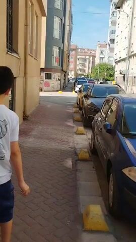 A person walks down a paved alleyway lined with parked cars. The alley is flanked by buildings on both sides. The person is wearing a white shirt and blue pants. The cars are parked along the curb, and yellow parking blocks are visible. The sky is blue and clear, indicating a sunny day. The buildings appear to be apartment buildings. The alleyway is relatively narrow.
