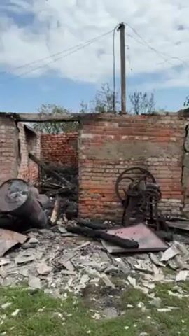 The video shows the remains of a brick building that has been severely damaged by fire. The structure is largely reduced to its brick walls, with a roof that is significantly damaged and partially missing. Debris, including charred wood, metal, and other unidentifiable materials, is scattered across the ground in front of the building. Two large metal barrels are visible among the wreckage. The sky is blue with scattered clouds. An electrical pole with wires is also visible near the building. The setting suggests a rural or suburban environment.
