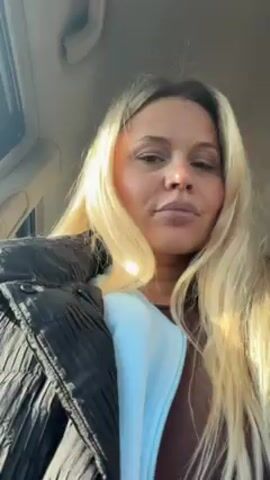 A woman with blonde hair is recording a video of herself inside a car. She is wearing a brown top, a light blue jacket, and a black puffer jacket. She looks directly at the camera with a neutral expression. The car appears to be in motion, and the sun is shining through the windshield.