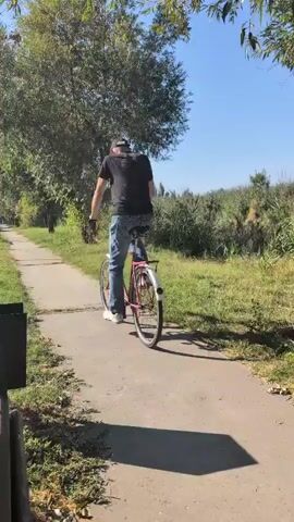 A person wearing a black t-shirt and blue jeans is riding a bicycle on a paved path. The path is surrounded by grass and trees, with a large tree prominently featured behind the cyclist. The sky is blue and clear, suggesting a sunny day. In the foreground, a park bench and a trash can are visible, casting shadows on the path. The cyclist is moving away from the camera.