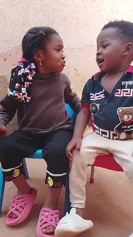 Two young children, a girl and a boy, are sitting on small plastic chairs. The girl is wearing a brown turtleneck shirt, black pants, and pink sandals. Her hair is braided with colorful beads. The boy is wearing a blue polo shirt with a teddy bear design and white pants. They appear to be talking and gesturing at each other. The girl then stands up, looks around, and walks away. The background is a simple outdoor setting with a partially visible wall and a metal door. The ground appears to be a dirt or sand surface.