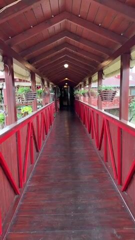 A first-person perspective video shows walking down a long wooden walkway. The walkway is covered by a wooden roof and has red railings on both sides. The railings have a decorative pattern of red beams. Hanging baskets with plants are placed along the sides of the walkway. The walkway is illuminated by lights embedded in the ceiling. The floor of the walkway is made of dark red wooden planks. The walkway leads to a distant dark opening.
