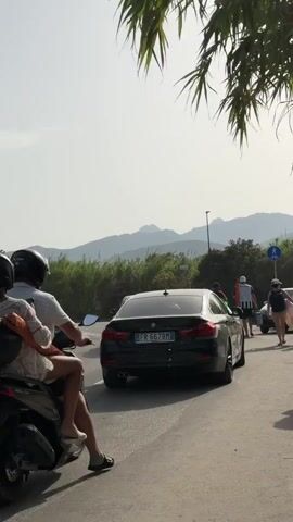 A scooter with two passengers wearing helmets is stopped in traffic. The scooter is facing away from the camera. The passengers are a man and a woman. The woman is sitting behind the man and is wearing a light-colored dress. The man is wearing a white shirt and shorts. In front of the scooter is a black car, also stopped in traffic. Behind the car, there are several other cars and people walking. The scene appears to be on a road with mountains in the background. There are trees and foliage on either side of the road. The sky is clear and bright.