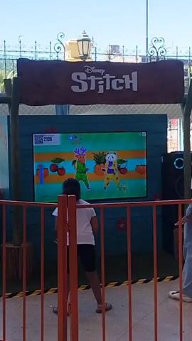The video shows two people, likely a mother and child, dancing in front of a screen displaying a "Disney Stitch" themed game. They are separated from the screen by an orange fence. The child is on the left wearing a white shirt and black shorts, actively dancing to the game's instructions, while the mother stands on the right, casually mimicking some moves. The screen shows animated characters dancing against a backdrop of fruits, and a score is visible in the corner. In the background, there are lampposts, vegetation, and glimpses of other people moving around. The area seems to be outdoors under daylight.