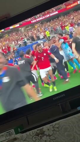 The video shows a group of male soccer players in red jerseys celebrating on a soccer field. The players are smiling and hugging each other. In the background, there are spectators in the stands. A large screen in the foreground shows the soccer match. The text on the screen reads "FIFA WORLD CUP 2026". The players are celebrating a victory and likely advancing to the next round of the World Cup.