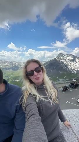 A woman with long blonde hair and sunglasses is taking a selfie video in a mountainous landscape. She is wearing a gray hoodie and gray sweatpants. Behind her, a parking lot is filled with motorcycles and a few cars. The background features a winding road, green hills, and snow-capped mountains under a blue sky with white clouds. Another person's shoulder is visible on the left side of the frame.
