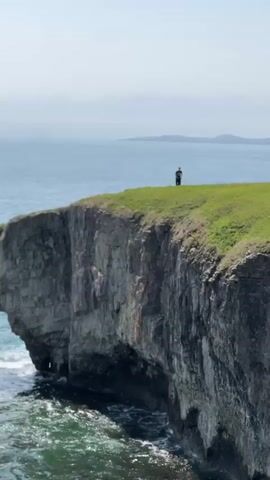 The video shows a rocky cliff with an archway carved into it by the ocean. A person stands on top of the cliff, briefly visible, then walks away. The camera focuses on the archway and the ocean waves crashing against the rocks. The sky is overcast.
