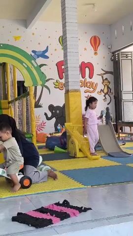 A young child in a light green t-shirt and shorts is on a white and orange ride-on toy. A woman wearing a black hijab and gray pants is sitting behind the child, holding onto the toy. The child is looking to the left. To the right of them, a young girl in pink pajamas stands on a yellow slide. She is holding a small blue object in her right hand. The environment appears to be an indoor play area with colorful wall murals depicting animals and hot air balloons. There are soft blue and yellow mats on the floor. In the background, a slide is visible, and a doorway leads to another area. The overall scene is well-lit.