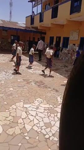 The video captures an outdoor scene at what appears to be a school or community center in Badiko, Nigeria, under bright, sunny daylight. The setting features a paved courtyard with irregular light-colored tiles, surrounded by yellow-painted buildings with blue window frames. One building has a metal-railed balcony, and another has a corrugated metal roof. A whiteboard with unreadable text is visible on one of the yellow walls.

The primary subjects are a group of young boys, likely primary school-aged, dressed in light-colored short-sleeved shirts and dark shorts, possibly school uniforms. Some boys are barefoot, while others wear shoes or socks. An adult male is briefly seen in the background.

The boys are energetically engaged in a game that resembles a form of tug-of-war or a group pulling/pushing activity, possibly using a rope or string (though the object being pulled is not always clearly visible). They are dynamically moving, pulling each other, and shouting with excitement. Their expressions and actions convey a sense of fun and active play. The sounds of their shouts and laughter further emphasize their energetic engagement in the game.