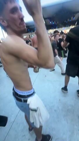 A young man with short dark hair and a mullet, shirtless and wearing jeans with his boxers visible, dances in an outdoor setting. He is in the foreground, facing away from the camera and to the right, with his right arm bent and his left arm gesturing. He appears to be at a crowded event with other people dancing around him. In the background, there is a stage with lighting structures and more people. The ground is a light-colored surface, possibly concrete. The sky is blue and clear.