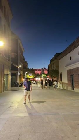 The video begins with a wide shot of a street at dusk or night. A man in a navy blue polo shirt and light-colored shorts walks towards the camera, his face obscured by a dark baseball cap. He is wearing white sneakers. The street is paved with interlocking stones and lined with buildings on either side. Streetlights illuminate the scene, casting long shadows. In the background, other people can be seen walking and congregating, and there are indications of outdoor dining or seating areas, with tables and chairs visible. Buildings in the background appear to be multi-story and of mixed architectural styles. The sky is a deep blue, indicating the transition from day to night. As the video progresses, the man continues to walk, and the camera remains focused on him. He gestures with his hands as he walks. The perspective shifts slightly as he moves past. The scene appears to be in an urban or town setting, possibly a pedestrianized area or a street with limited vehicle traffic.