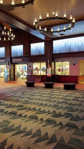 The video shows the interior of a large lobby or common area with a patterned carpet and a large circular chandelier with lights. In the background, there are storefronts with signage that reads "Emporium" and "Candy Shop." A hallway extends into the distance with people walking. The walls are decorated with images of trees. There are several dark, cylindrical benches in the foreground.