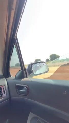 The camera is inside a car, focusing on the driver's side door and side mirror. The car is traveling on a dirt road under a sunny, partly cloudy sky. The cracked front windshield is partially visible, and a few trees are visible in the background. The interior of the car appears dark in contrast to the bright exterior.