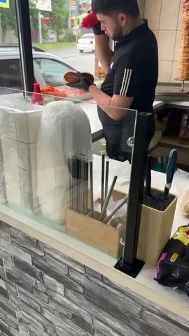 A man in black is preparing food behind a counter with a glass screen. He is wearing black gloves and adding a red sauce from a squeeze bottle into a sandwich. Behind him is a vertical skewer of meat. The counter has a variety of items, including plastic-wrapped containers, a wooden holder with straws and utensils, and a knife block. The counter is made of grey brick-like material. Outside the window, cars are visible on a street.
