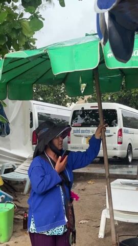 A woman stands under a green umbrella in what appears to be a rainy outdoor setting. She is wearing a blue jacket, a patterned skirt, a hat, and a face mask. She is holding a smartphone to her face as if she is talking on it. She is also holding onto the pole of the umbrella for support. Behind her, there are two white vans. The ground appears wet and sandy. To her left is a green bucket, a bottle, and some beach chairs. To her right is a white cooler.