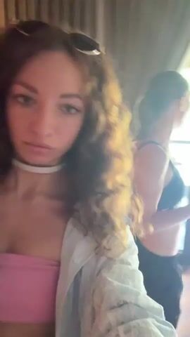A video shows two women indoors. The woman in the foreground has long, curly brown hair, sunglasses on her head, a pink tube top, and a white lace-patterned overshirt. She looks towards the camera, then briefly turns away before smiling back at the camera. In the background, a second woman with shorter, light-colored hair is wearing a black Nike sports bra and black pants. She is seen adjusting her bra, then puts a small white object into her mouth, and subsequently raises her right arm and moves her body. The background features curtains and a window or door revealing an outdoor view with greenery.