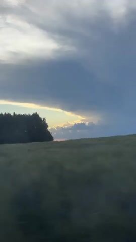 The video shows a blurry landscape with a field in the foreground and a dark line of trees in the background. The sky is filled with dramatic, dark clouds, but a sliver of sunlight is visible peeking through a gap in the clouds above the trees. The overall impression is of a moody, overcast sky with a hint of light breaking through. The focus is soft, giving the impression of movement, perhaps from a car window.
