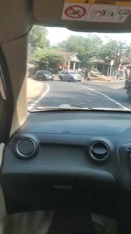 The video shows the interior of a car, looking out the windshield. On the road ahead, there are several cars. One car is a black SUV, possibly a Toyota Fortuner, and another appears to be a silver SUV. There are also trees and houses visible in the background. The sun appears to be shining. There are also some warning stickers on the car's visor, including one that depicts a person being thrown from a vehicle.