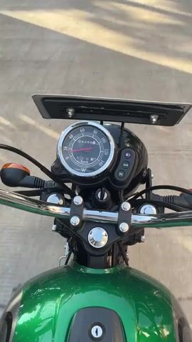 The video shows a first-person perspective from a Kawasaki W175 motorcycle. The motorcycle is green with chrome handlebars and has an analog speedometer showing a reading of 4266 km. The bike is idling, and the view is from the rider's seat looking down a narrow, paved road between houses with trees lining either side. The sky is visible through the trees, indicating a daytime setting. There is no visible rider or any interaction with people.