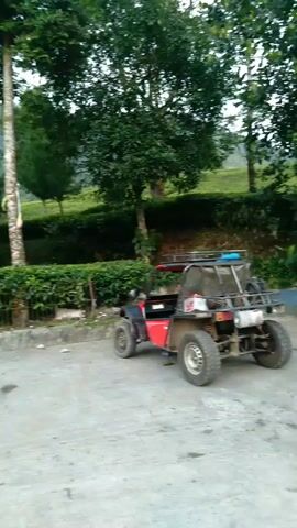 The video shows several ATVs parked in a lot near some greenery. The ATVs are various colors including black, red, and green. A silver minivan arrives, and several men get out and stand near it. There is a banner in the background, and a forested mountain looms in the distance. The sky is overcast.