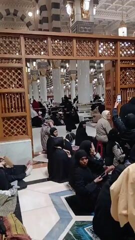 The video shows the interior of a mosque. Many people, mostly women, are sitting on the floor in groups. Most of the women are wearing black abayas and headscarves. Some people are using their cell phones. A woman on the right side of the frame is taking a picture with her phone.  The mosque has tall white pillars with black and white striped arches above. There are also ornate wooden screens separating different areas. The floor has a geometric pattern in blue, white, and gray. Chandeliers hang from the ceiling. The scene appears to be a break period for the visitors.