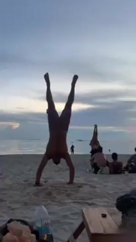 The video shows a beach scene at sunset. In the foreground, a shirtless man in shorts is doing a handstand on the sand. Further away, another person is also doing a handstand. Several people are sitting or lying on the beach, watching the sunset, and some are standing near the water. In the foreground, there's a water bottle, a bag, and a wooden surface, possibly part of a chair. The sky is cloudy with the colors of the sunset peeking through.