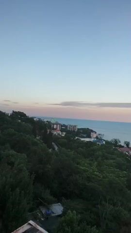 A wide aerial view of a coastal town at dusk shows buildings nestled among lush green trees. The sea stretches out to the horizon, meeting a pale pink and blue sky. The camera slowly pans to the right, revealing more of the town and the vast expanse of water.