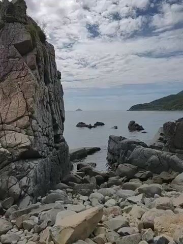 A wide shot shows a rocky coastline with the ocean in the background. Large, weathered rocks and boulders litter the foreground, leading down to the water's edge. In the distance, the ocean stretches to the horizon, with a few smaller rocks visible just above the surface. A hazy, mountainous landmass can be seen on the right side of the frame, and a small, distant island is visible on the left. The sky above is filled with scattered, fluffy clouds. The lighting suggests it is daytime, possibly overcast or late afternoon. There are no people or animals visible in the scene. The overall impression is one of a rugged and natural coastal landscape.