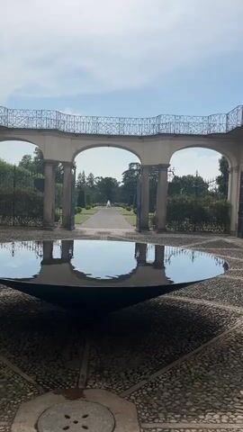 A very still, wide shot shows a reflective fountain in the foreground. The fountain has a dark, conical basin. Behind the fountain, a walkway extends into a garden. The walkway is flanked by manicured hedges and trees. In the distance, a fountain can be seen at the end of the walkway. The walkway is framed by a colonnade with three arches and a decorative railing. The sky above is blue with scattered clouds.