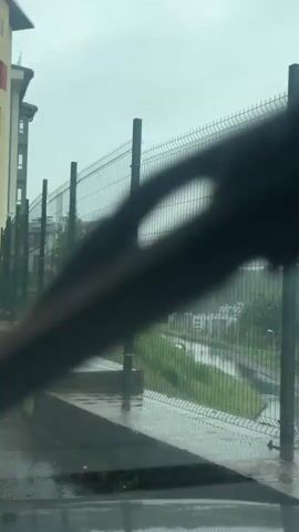 The video shows a wet, overcast day in Близкое Кп, Russia. The scene is dominated by a tall, green wire mesh fence that stretches across the frame, supported by dark metal posts. The fence appears to be surrounding an area, with a building on the left side of the image and a treeline with some houses visible in the distance beyond the fence.

The ground in the foreground is wet and reflective, indicating it has been raining or is currently raining. Water pools on a concrete structure that runs alongside the fence. The weather is gloomy, with a thick, light grey sky that suggests it is either early morning, late afternoon, or simply a very cloudy day. There are no people visible in the frame. The overall mood is somber and quiet, reflecting the muted colors and the absence of activity. The focus is on the texture of the wet surfaces and the repetitive pattern of the fence.
